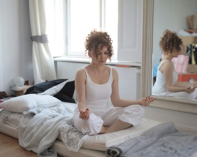 Tranquil female in sleepwear sitting on bed in messy room and practicing yoga with closed eyes while maintaining mental health