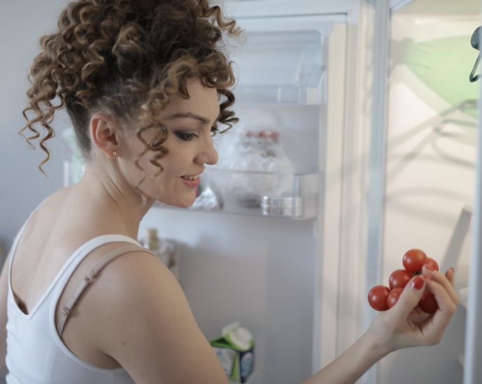Side view of content female in casual wear standing in kitchen near opened fridge and picking fruit for breakfast