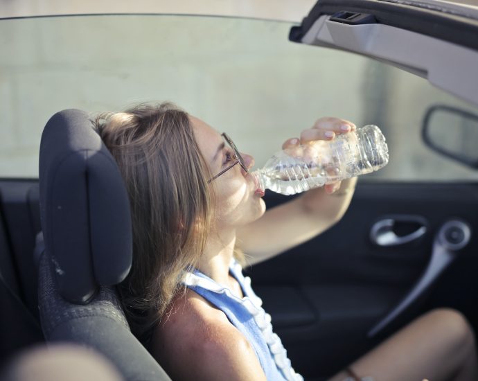 High angle side view of young woman in glasses and casual clothes drinking water from plastic transparent bottle while sitting in cabriolet with open roof in traffic jam in hot sunny day