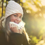 Young content female wearing warm hat and scarf standing in city garden and enjoying weekend while smiling and looking at camera