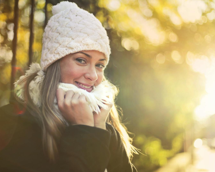 Young content female wearing warm hat and scarf standing in city garden and enjoying weekend while smiling and looking at camera