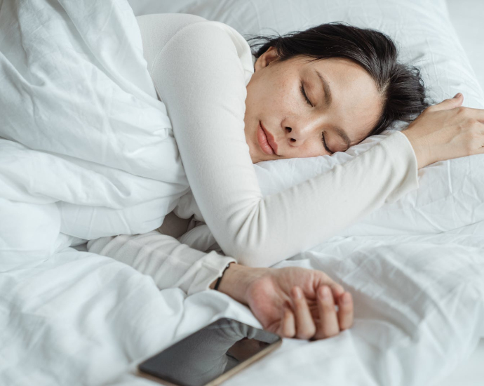 Calm Asian female wearing white pajama sleeping in comfortable bed with white sheets near modern mobile phone in morning