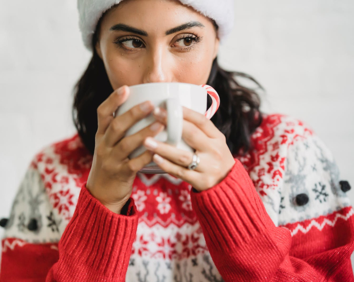 Young lady in Santa hat and sweater enjoying cup of cacao with candy cane in light room and looking away