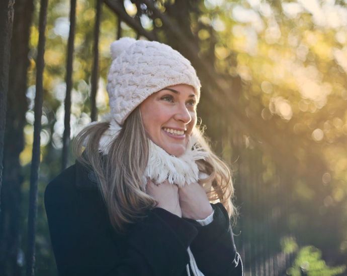 Photography of Woman in Black Jacket and White Knit Cap Smiling Next to Black Metal Fence