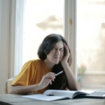 Ethnic female student in casual wear sitting at table with laptop and notebook and grimacing from migraine while doing homework during distance learning