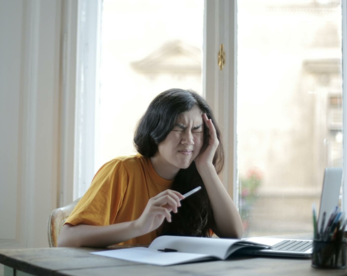 Ethnic female student in casual wear sitting at table with laptop and notebook and grimacing from migraine while doing homework during distance learning