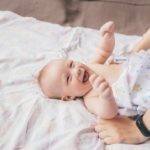 a smiling baby laying on a bed with a woman's arm