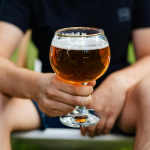 A man holding a glass of beer in an outdoor setting during the day.