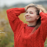 Woman in a red sweater smiling outdoors, enjoying a sunny day in nature.