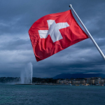 a swiss flag blowing in the wind near a body of water