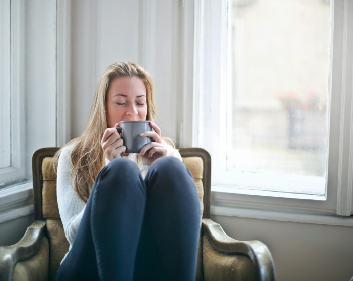 Woman enjoying a warm drink in a cozy armchair by a window, embracing relaxation.