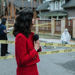 Female journalist reporting live on a crime scene investigation with police presence outdoors.