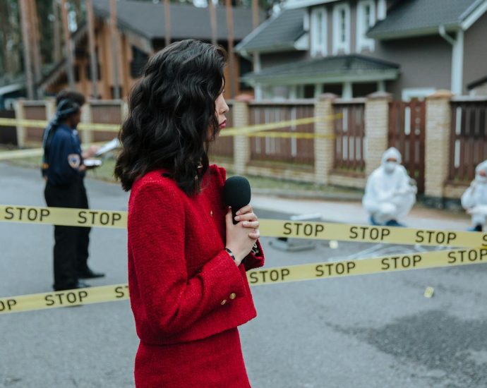 Female journalist reporting live on a crime scene investigation with police presence outdoors.