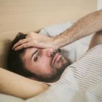 From above of sleepy bearded man just awakened rubbing eyes while lying on bed at home