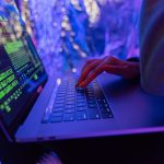 Close-up of hands typing on a laptop displaying cybersecurity graphics, illuminated by purple light.
