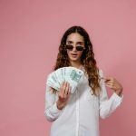 Woman in sunglasses holding and pointing at a stack of 100 euro notes against a pink background.