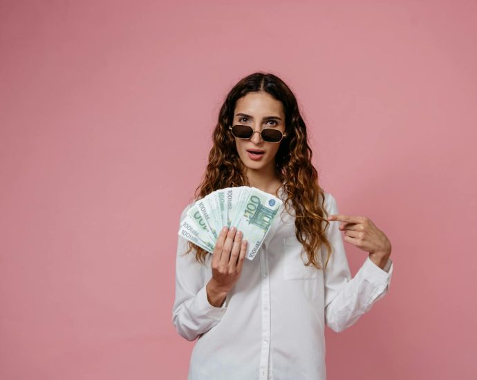 Woman in sunglasses holding and pointing at a stack of 100 euro notes against a pink background.