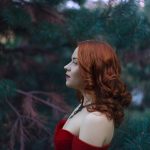 A red-haired woman poses in front of trees.