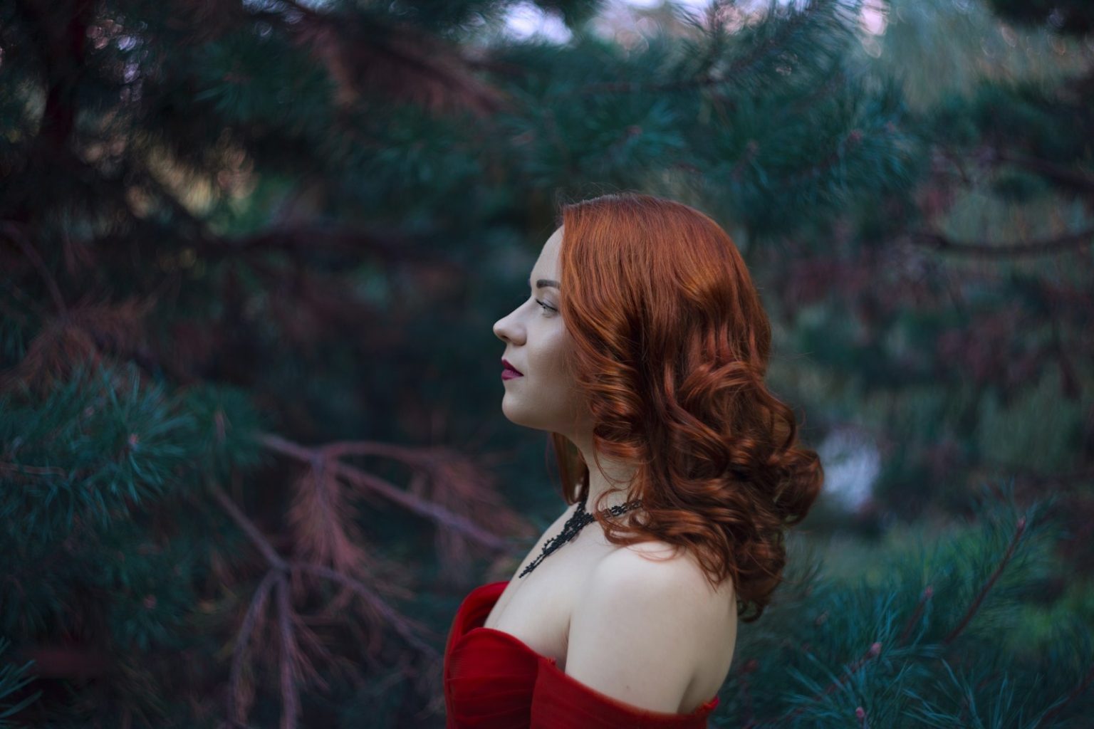 A red-haired woman poses in front of trees.