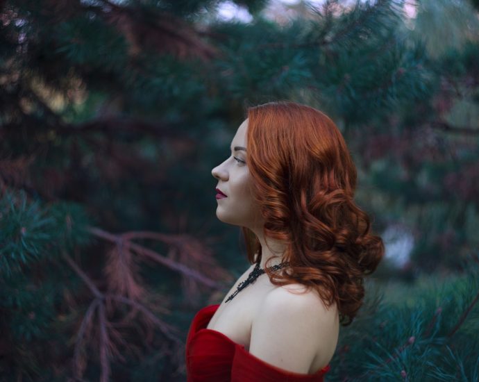 A red-haired woman poses in front of trees.