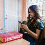 A teenage girl sits in a classroom, focused on her smartphone among books and educational materials.