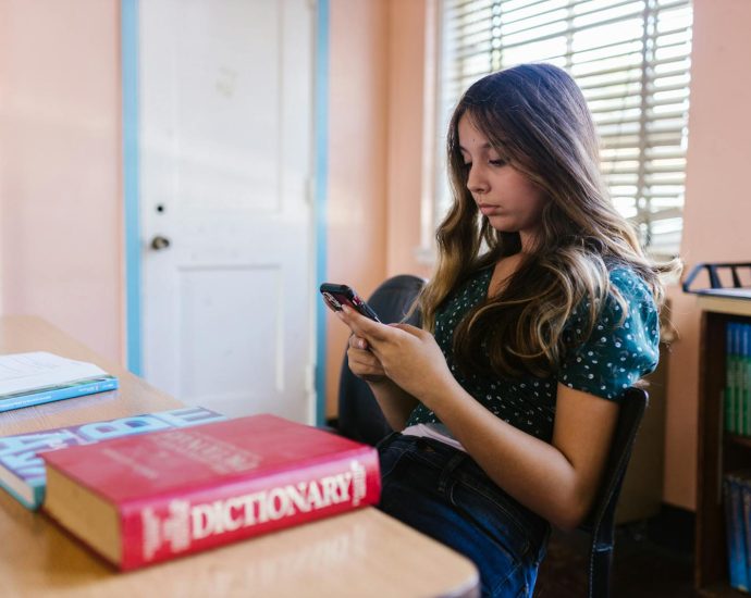 A teenage girl sits in a classroom, focused on her smartphone among books and educational materials.
