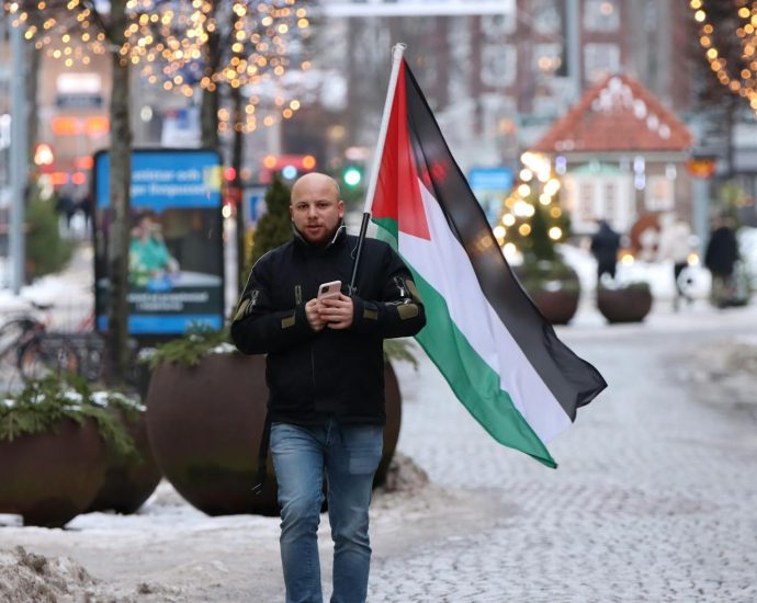 A man walks in Jönköping, Sweden, holding a flag on a winter street with festive lights.