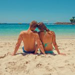 A couple enjoys a sunny day on the sandy seashore, surrounded by blue ocean and clear skies.
