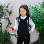 A young girl in school uniform smiling and holding a phone outdoors.