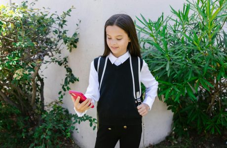 A young girl in school uniform smiling and holding a phone outdoors.