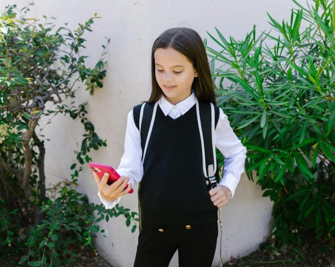 A young girl in school uniform smiling and holding a phone outdoors.