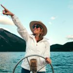 Smiling woman in sun hat and sunglasses pointing while steering a boat on Lake Minnewanka, Canada.