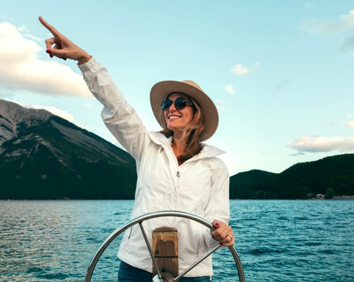 Smiling woman in sun hat and sunglasses pointing while steering a boat on Lake Minnewanka, Canada.