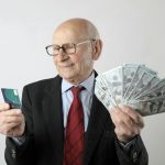 Elderly man in a suit holding a credit card and US dollar bills, representing finance and wealth.