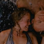 Joyful couple embracing beach waves, eyes closed, under summer sun.