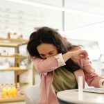 Woman in a cafe sneezing into her elbow while sitting at a table with a laptop.