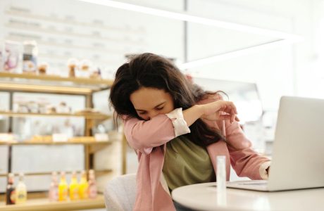 Woman in a cafe sneezing into her elbow while sitting at a table with a laptop.