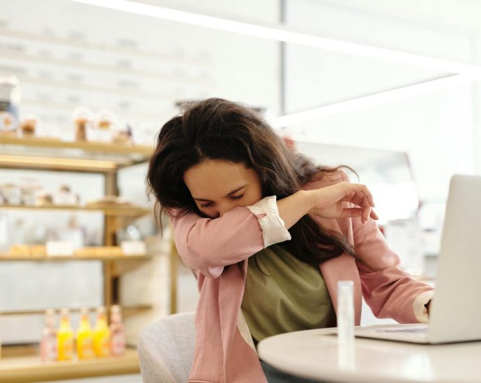 Woman in a cafe sneezing into her elbow while sitting at a table with a laptop.