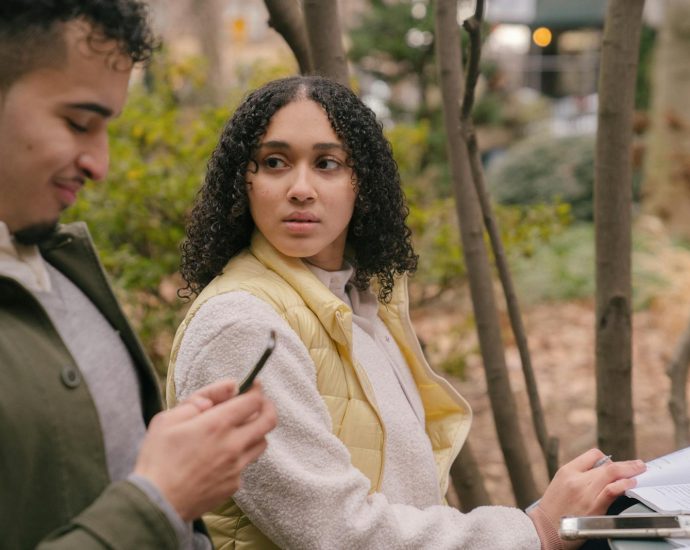 Side view of Hispanic female sitting at table with textbooks while preparing for exams with phone while looking jealously at man texting on cellphone in daytime in park