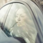 A woman reflects while sitting inside a car, looking through the window with a thoughtful expression.