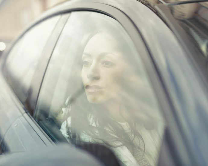 A woman reflects while sitting inside a car, looking through the window with a thoughtful expression.