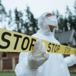 Investigator in protective gear securing crime scene with stop tape outdoors.