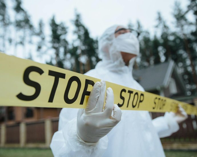 Investigator in protective gear securing crime scene with stop tape outdoors.