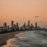 people standing on shore near city buildings and ocean during sunset