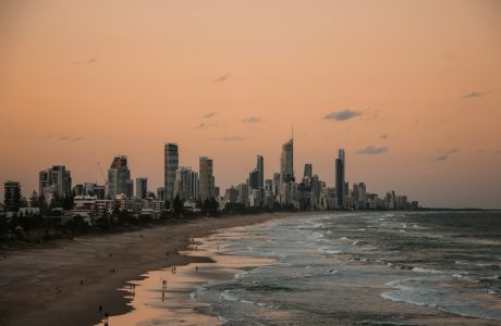 people standing on shore near city buildings and ocean during sunset