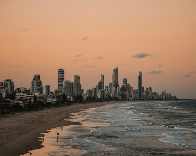 people standing on shore near city buildings and ocean during sunset