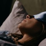 A serene scene of a woman wearing a blue eye mask, sleeping soundly in a cozy indoors setting.