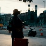 woman wearing coat standing on road with travel luggage during daytime