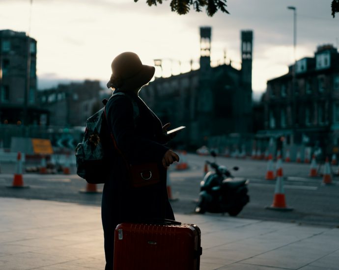 woman wearing coat standing on road with travel luggage during daytime
