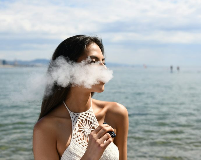 woman wearing white sleeveless top smoking tobacco while standing near blue sea under white and blue skies during daytime